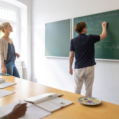 Diverse adults in an english language class: male teacher writes on chalkboard while female instructor stands by window as students listen and take notes in a bright classroom.