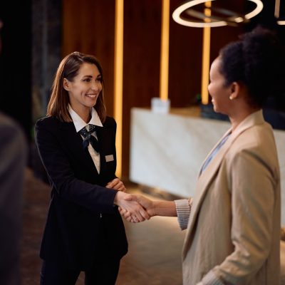 Happy hotel manager handshaking with her guests in a hallway.